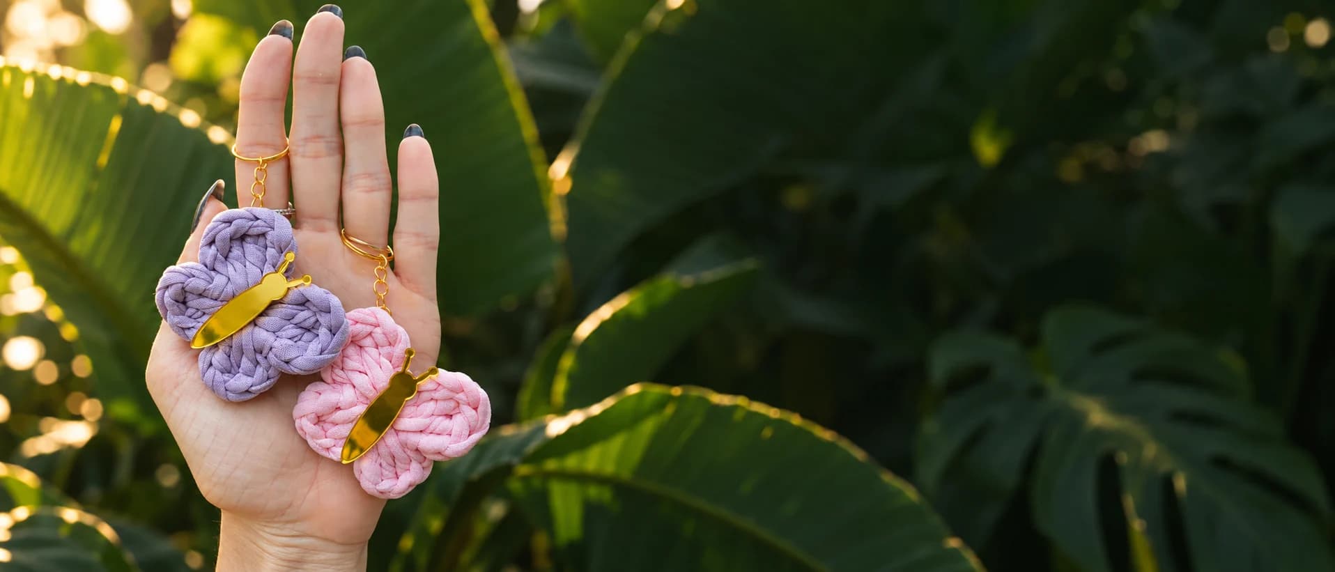 Mão aberta segurando dois chaveirinhos de borboleta em fio de malha, um lilás e um rosa, com pingentes dourados, na frente de folhas de bananeira ao pôr do sol
